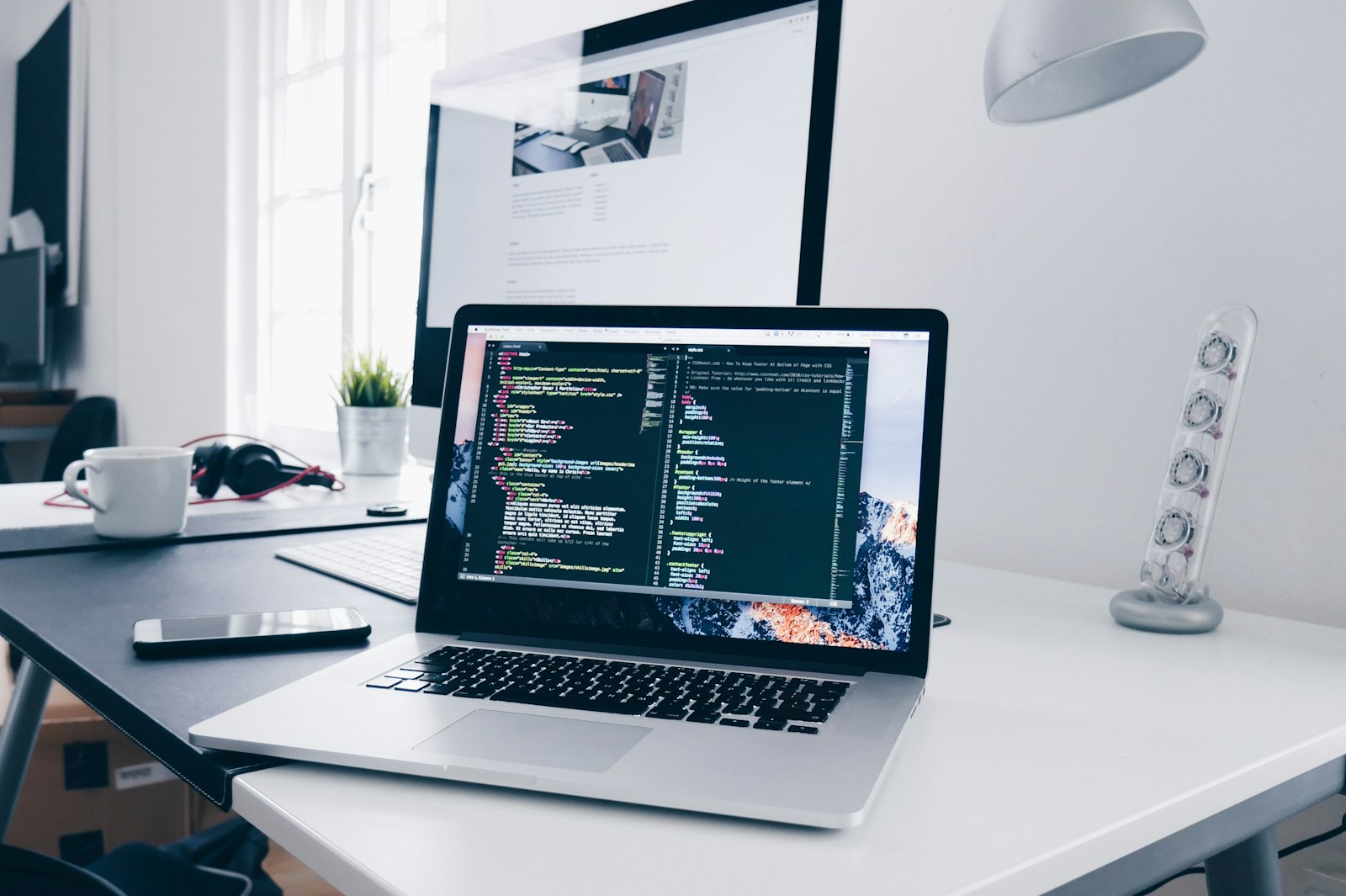 A laptop and monitor on a white desk showing source code