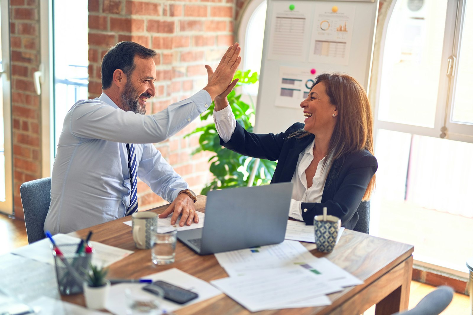 Two colleagues celebrating a result together across a desk in a bright office